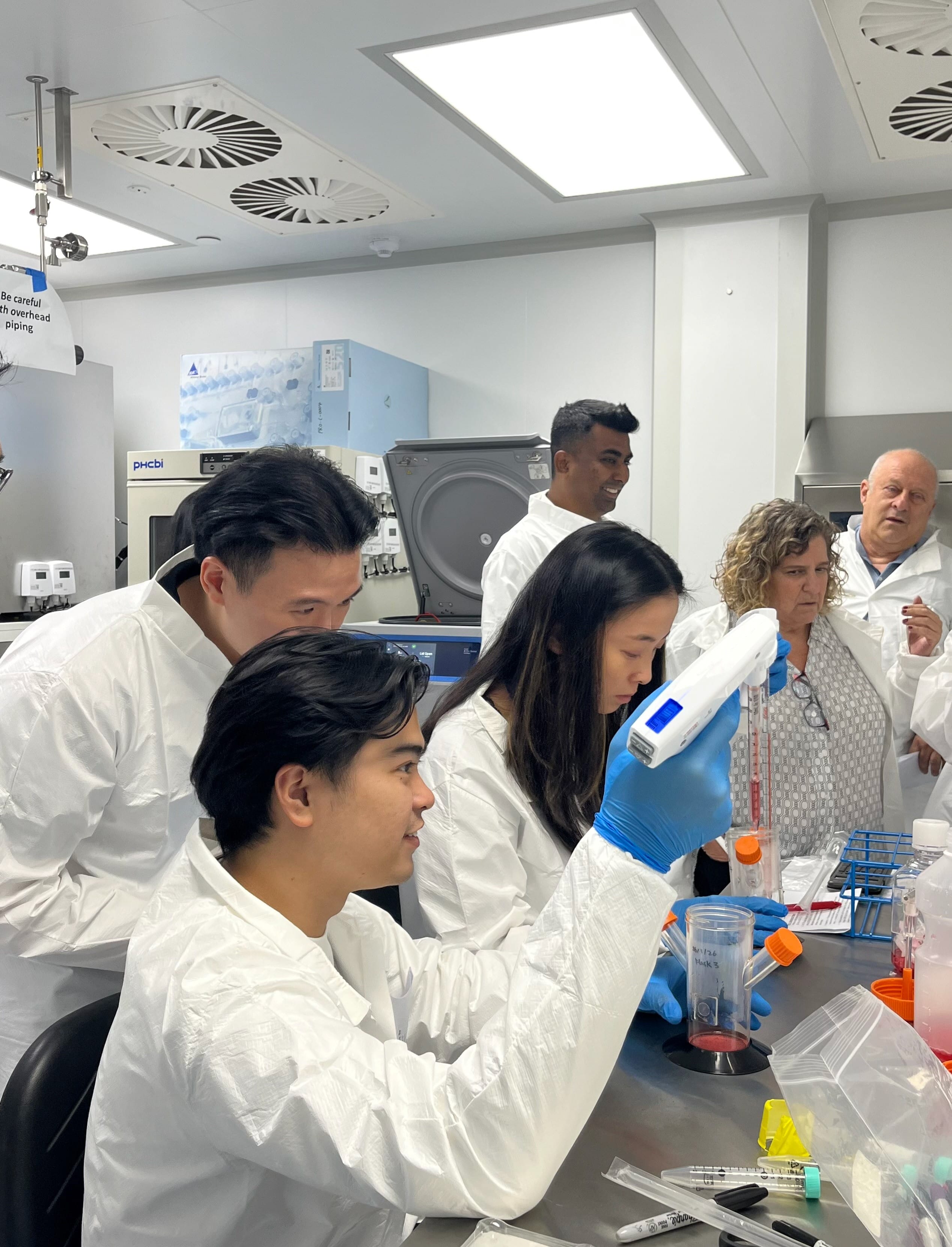 Participants in laboratory protective attire taking part in a hands-on cell culture training session, observing and performing laboratory procedures with equipment and sample containers in a research laboratory.